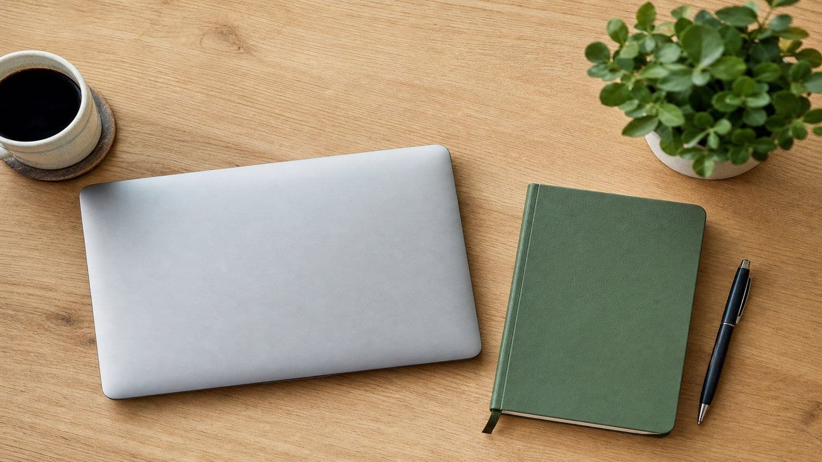 Neatly arranged home-office desk with a closed laptop, notebook, and small green plant seen from above