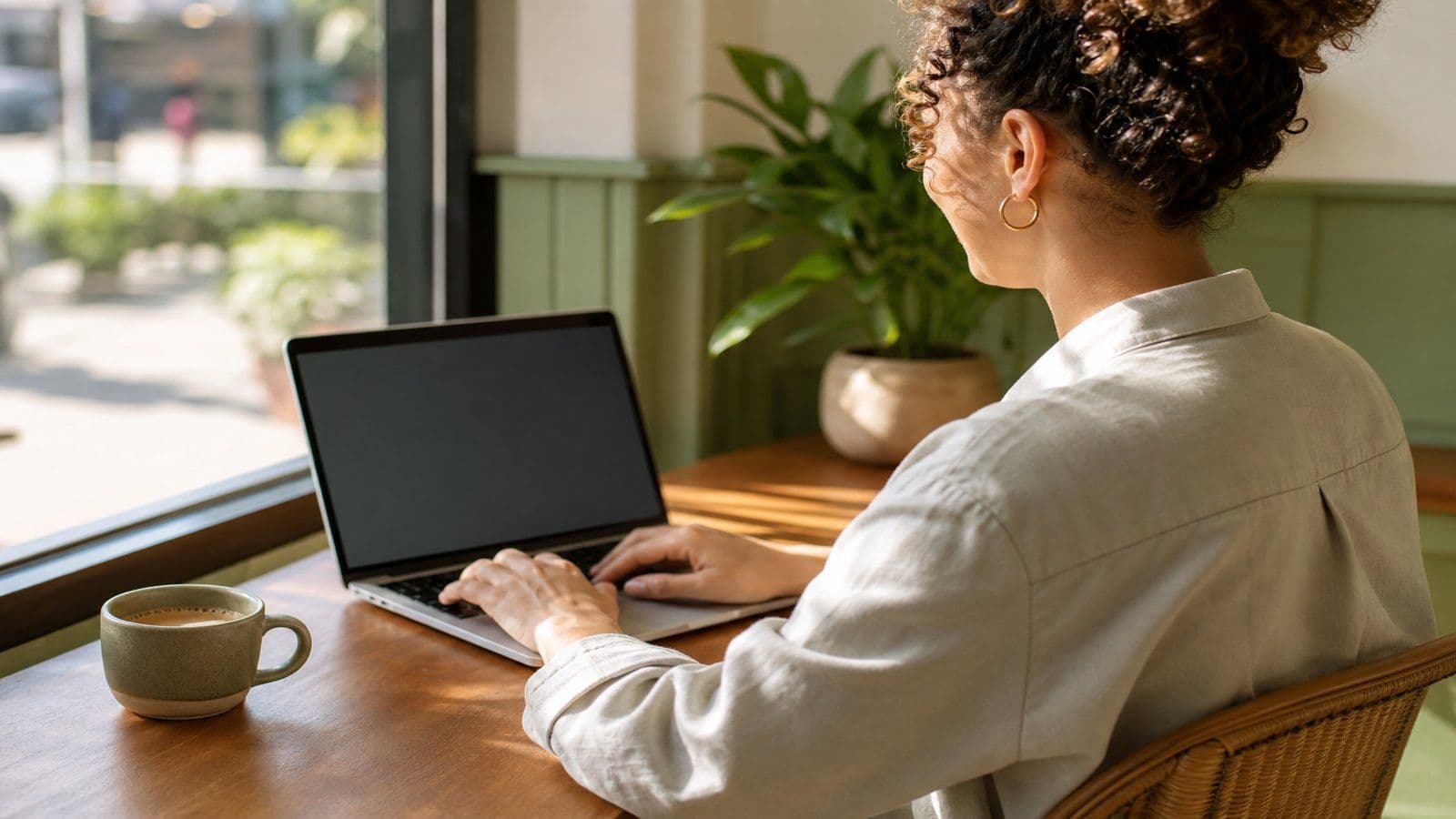Woman in business-casual working on a laptop in a sunny cafe corner seen over the shoulder